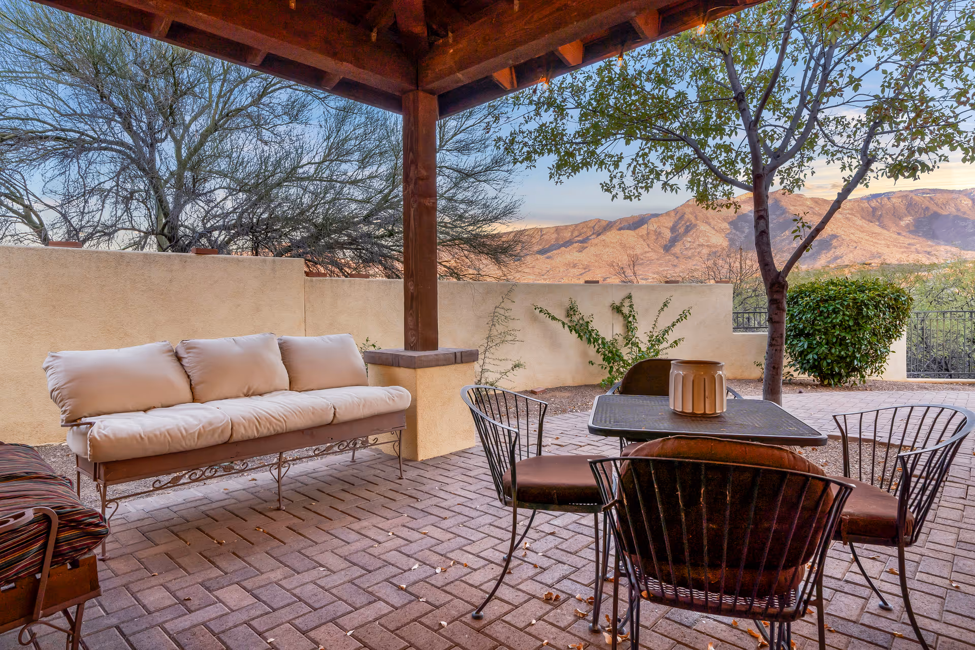 Outdoor patio area with a cushioned bench, metal table, and four metal chairs under a wooden pergola. The patio is paved with bricks and surrounded by a low beige wall, with trees and shrubs visible. In the background, there are mountains under a clear sky.