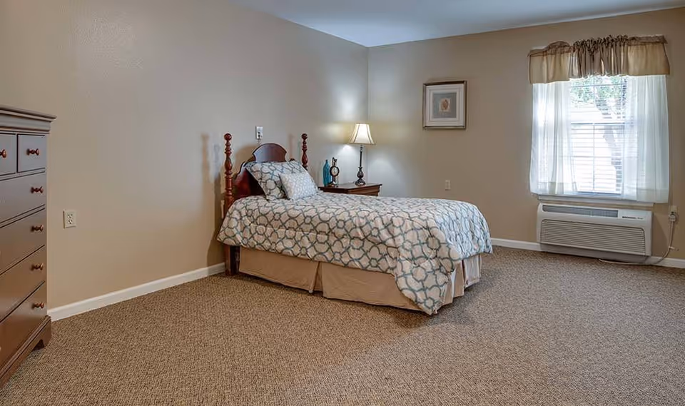 Furnished bedroom with a single bed, nightstand and lamp, dresser, and a window with an air-conditioning unit.