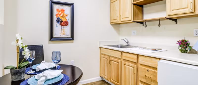A small kitchen area with light wood cabinets, a sink, and a white countertop. To the left, there is a round dining table set for two with blue placemats, white napkins, blue glasses, and a potted orchid plant. A framed floral painting hangs on the wall above the table.