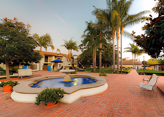 Outdoor courtyard area with a decorative water fountain in the center, surrounded by brick pathways, benches, potted plants, and tall palm trees. Residential buildings and shaded seating areas are visible in the background under a clear sky.