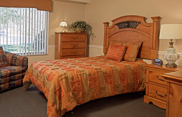 A cozy bedroom with a wooden bed frame and a bed covered with a patterned orange and beige quilt. There are two pillows on the bed. To the right of the bed is a wooden nightstand with a decorative lamp and a digital alarm clock. To the left of the bed is a wooden chest of drawers with a small lamp and a potted plant on top. A patterned armchair is positioned near a window with vertical blinds, letting in natural light.
