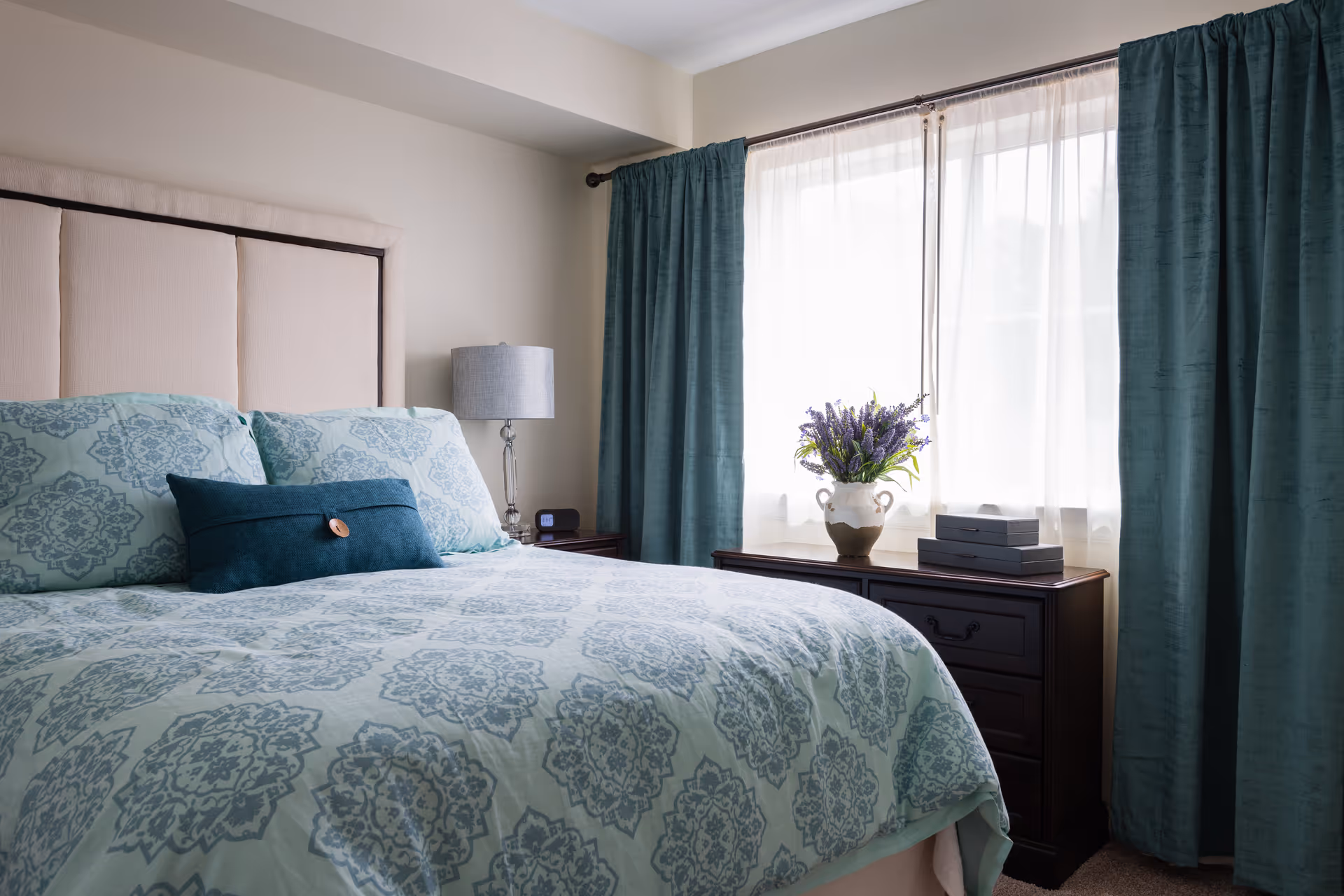 A bright bedroom featuring a bed with teal patterned bedding, an upholstered headboard, a nightstand with a lamp, and a dresser beneath a curtained window with a vase of flowers.