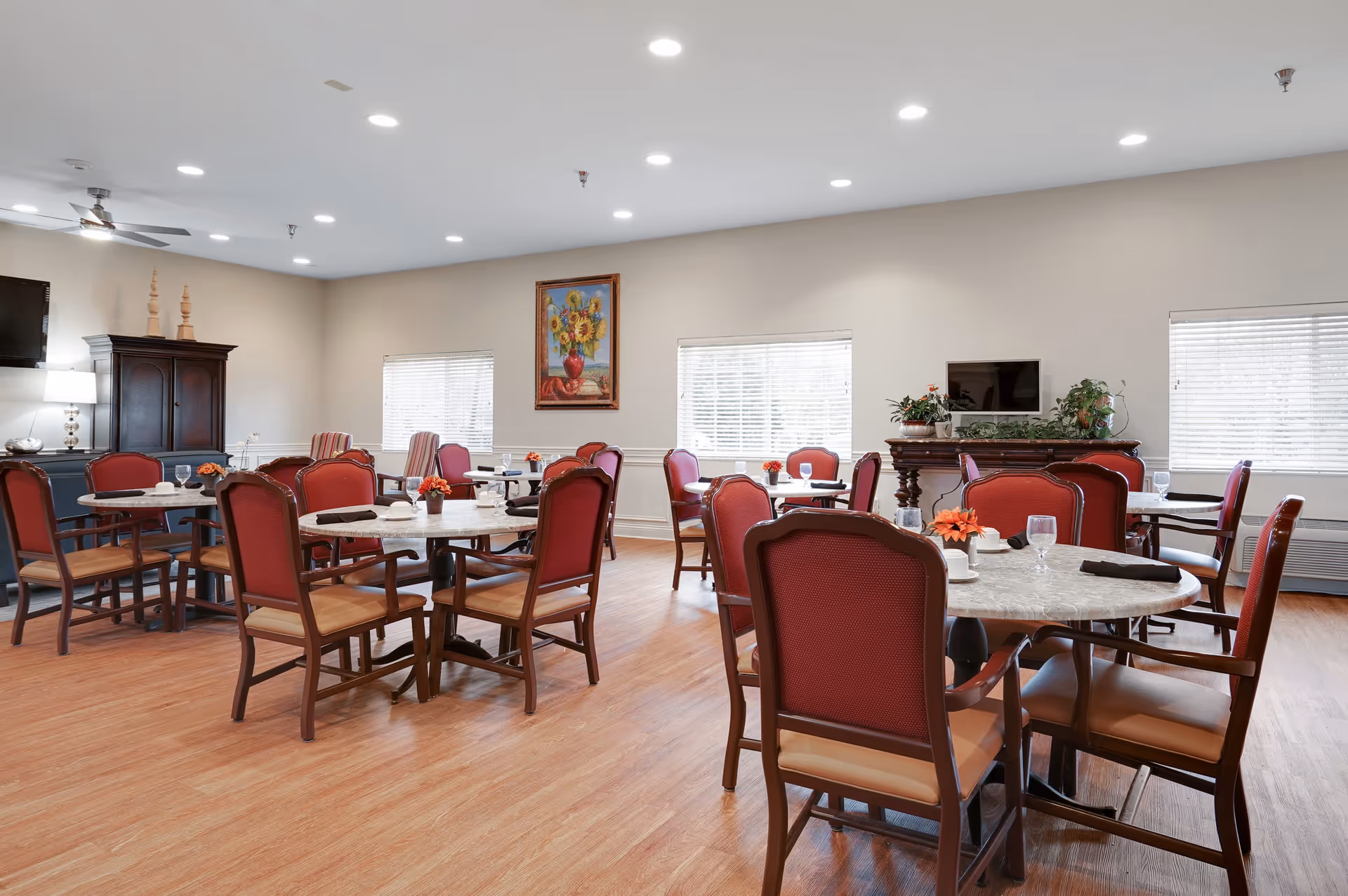 A bright and spacious dining room with multiple round tables surrounded by wooden chairs with red cushions. The room features wood flooring, beige walls, three windows with blinds, a painting of sunflowers on the wall, and a dark wooden cabinet and fireplace mantel decorated with plants and a small TV.