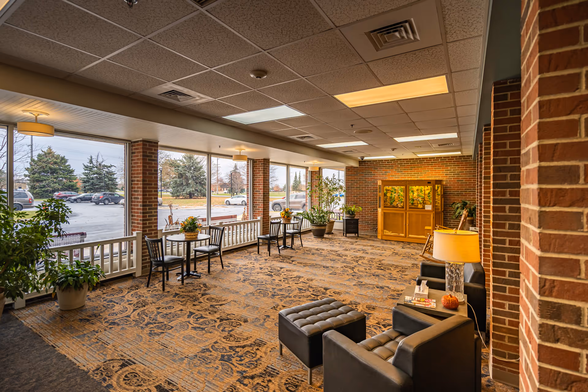 A bright lobby/waiting area with chairs, ottomans, small tables, potted plants and large windows looking out to a parking lot.