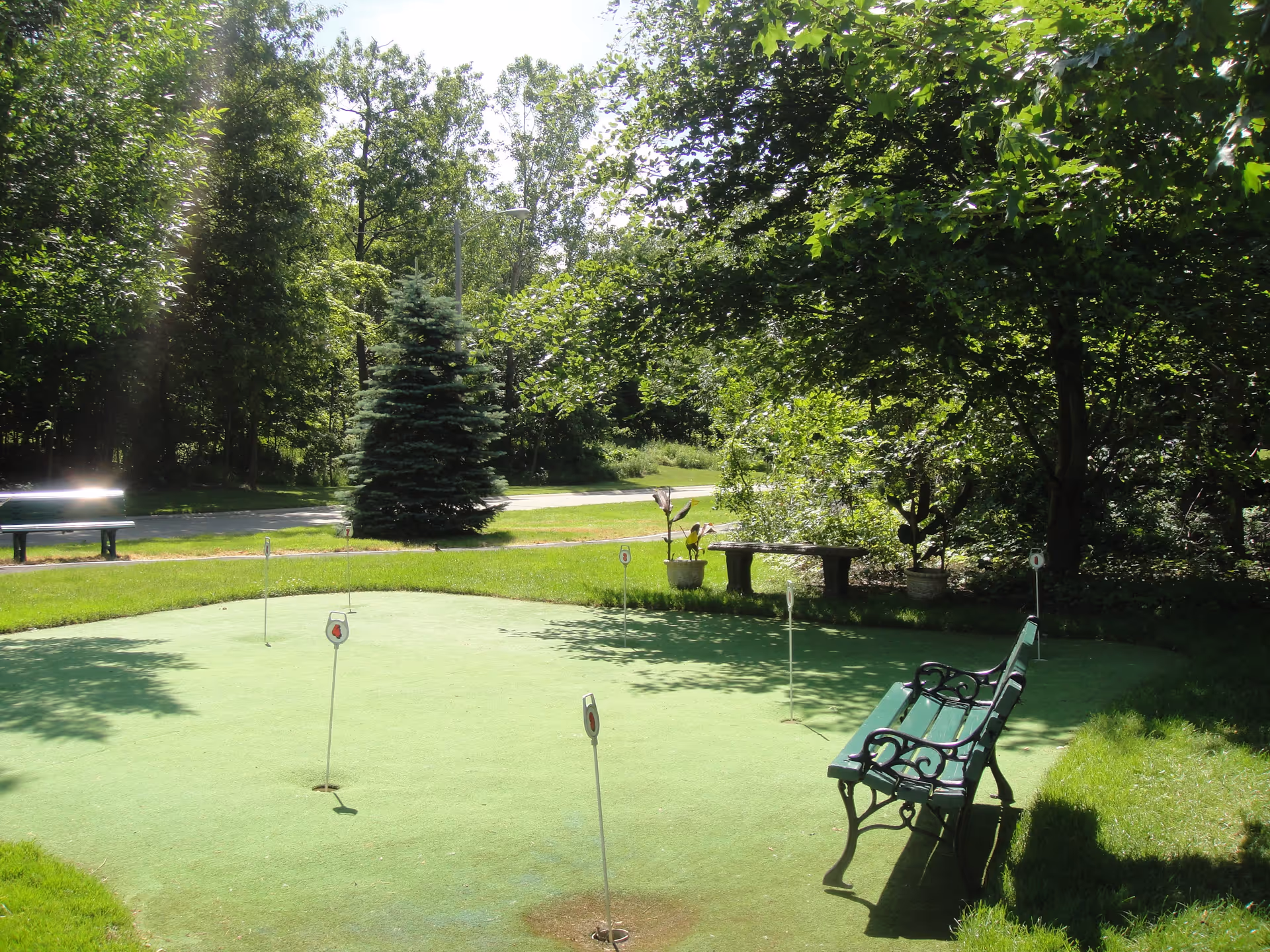 A small outdoor putting green surrounded by lush green grass and trees. There are several golf holes with flags on the putting green, a green bench with ornate black metal armrests, a stone bench, and potted plants around the area. The scene is sunlit with shadows from the trees.
