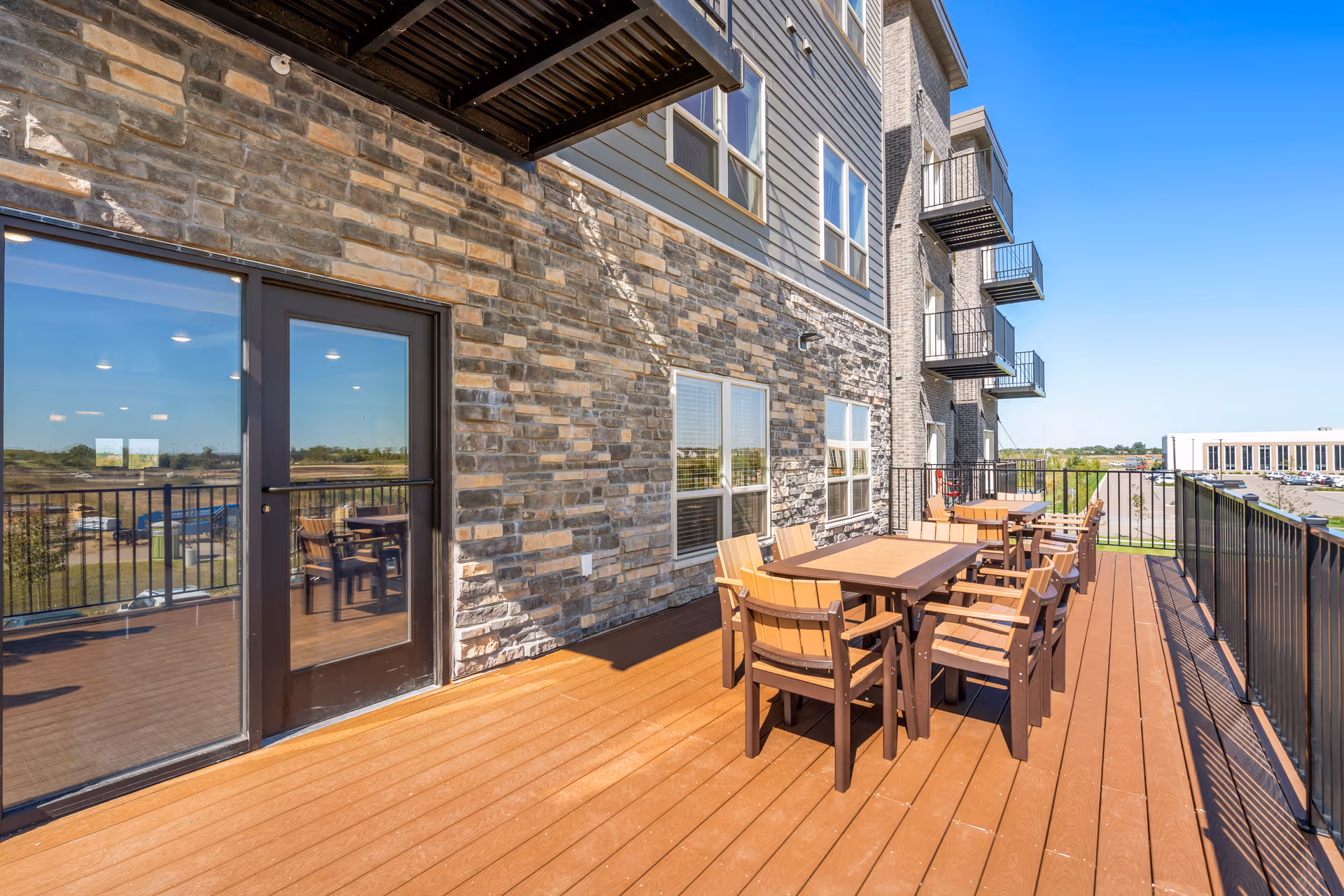 Raised outdoor deck with multiple wooden tables and chairs alongside a stone-clad senior living building under a clear blue sky.