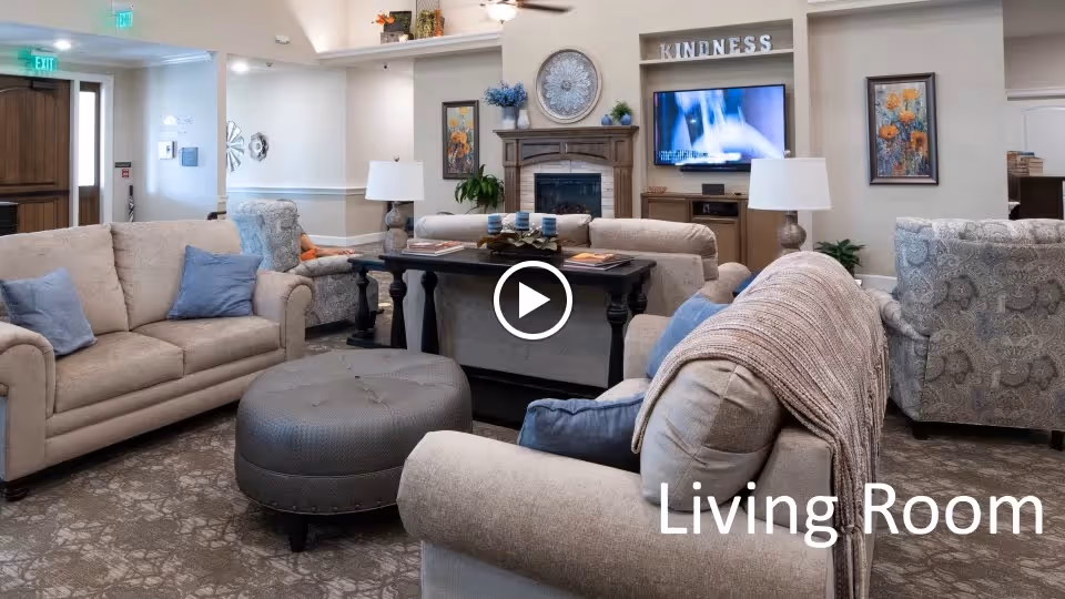 A cozy living room in an assisted living facility featuring beige sofas with blue and gray cushions, a round gray ottoman, a dark wooden coffee table with candles and books, a fireplace with decorative items above it, a wall-mounted TV, and framed artwork on the walls. The room has a carpeted floor and soft lighting.