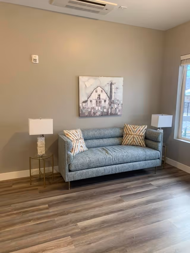 Small seating area with a blue upholstered sofa, two patterned pillows, side tables with lamps, and a framed painting on a beige wall.