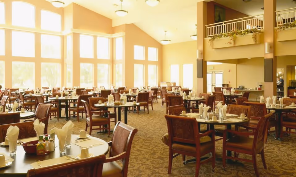 Spacious sunlit dining room with multiple round tables set for meals and large windows.