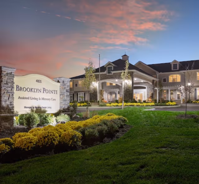 Exterior view of Brooklyn Pointe Assisted Living & Memory Care facility at dusk, showing a well-lit entrance with stone pillars, manicured landscaping with yellow flowers, and a sign displaying the facility's name.