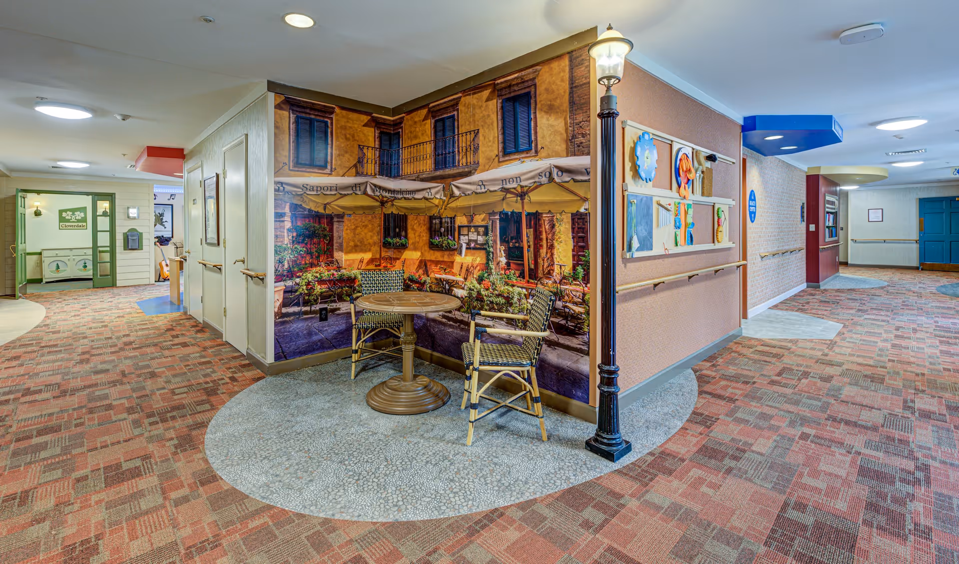 A brightly lit hallway in a senior living facility with patterned carpet and handrails along the walls. There is a small seating area with a round table and two chairs in front of a wall mural depicting an outdoor café scene. A decorative street lamp stands next to the seating area. The hallway has colorful ceiling accents and artwork on the walls.