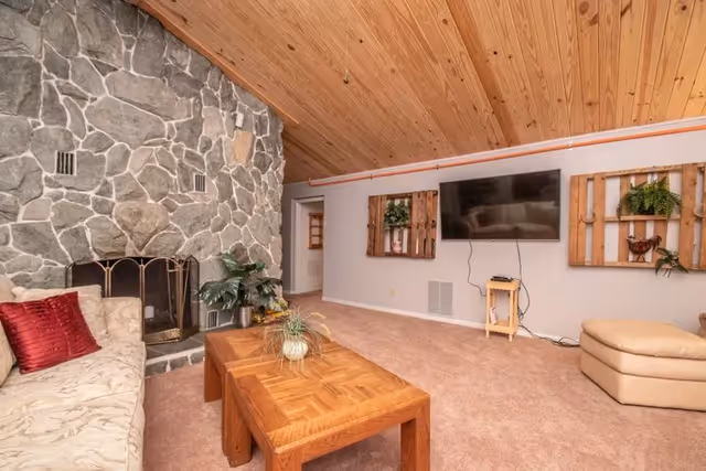Cozy living room with a stone fireplace, wood-paneled vaulted ceiling, wall-mounted TV, sofa, and wooden coffee table.