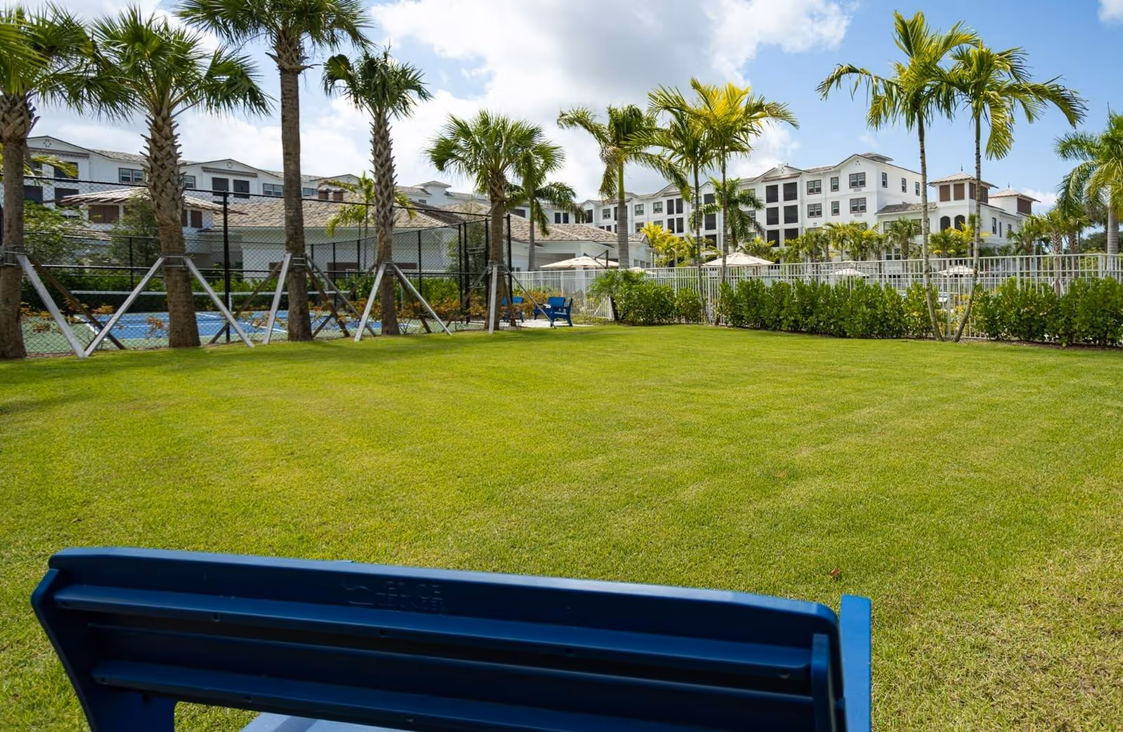 View of a well-maintained green lawn with palm trees and a blue bench in the foreground. In the background, there is a fenced area with a tennis or basketball court and a large multi-story white building under a partly cloudy sky.