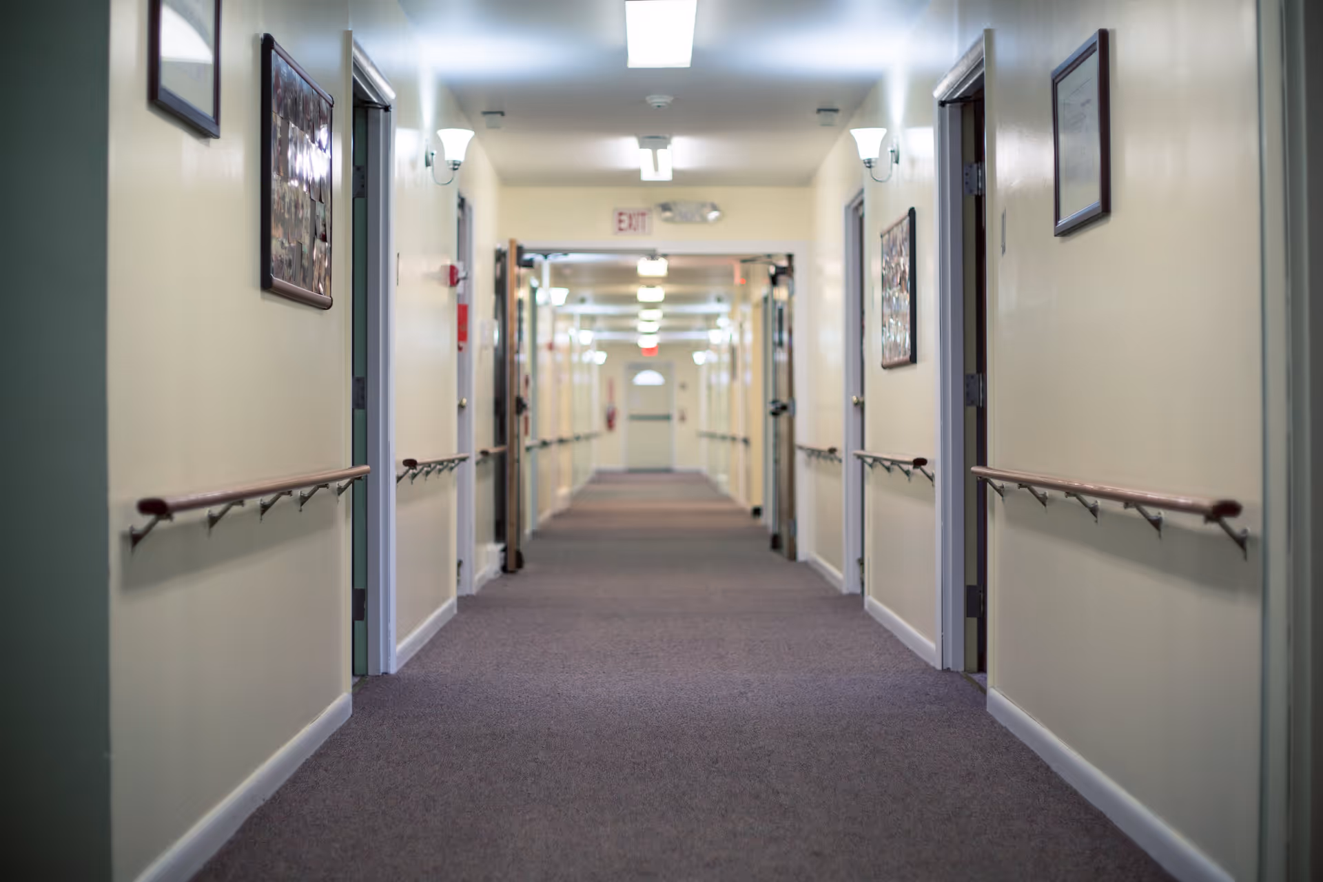 A long, carpeted hallway in a senior living facility with handrails on both sides, cream-colored walls, framed pictures hanging on the walls, and multiple open doors leading to rooms. The hallway is well-lit with wall-mounted lights and ceiling lights, and an exit sign is visible at the far end.