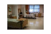 Shared resident room with two hospital-style beds, privacy curtains, bedside tables and a window with patterned curtains.