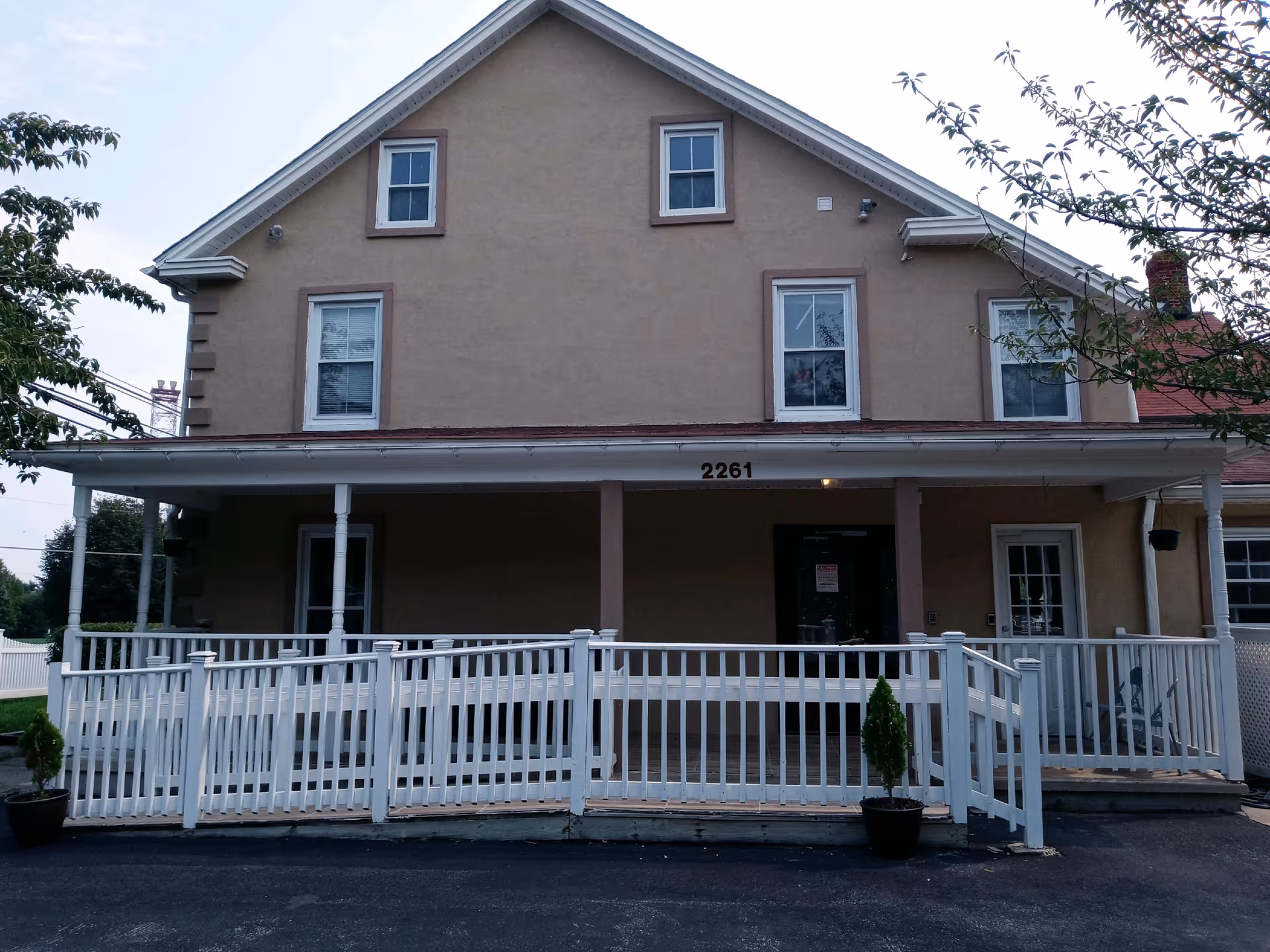 Front exterior of a two-story beige assisted living house with a covered porch, white railing, and the number 2261 above the entrance.