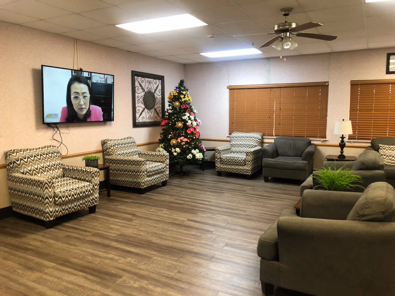 Communal lounge with multiple upholstered chairs, a decorated Christmas tree, a wall-mounted TV, and wood-look flooring.