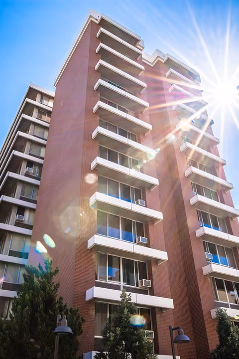 Exterior view of a tall brick apartment building with multiple balconies and windows, some with air conditioning units. The sun is shining brightly in the clear blue sky, creating lens flare effects. Trees and street lamps are visible at the base of the building.
