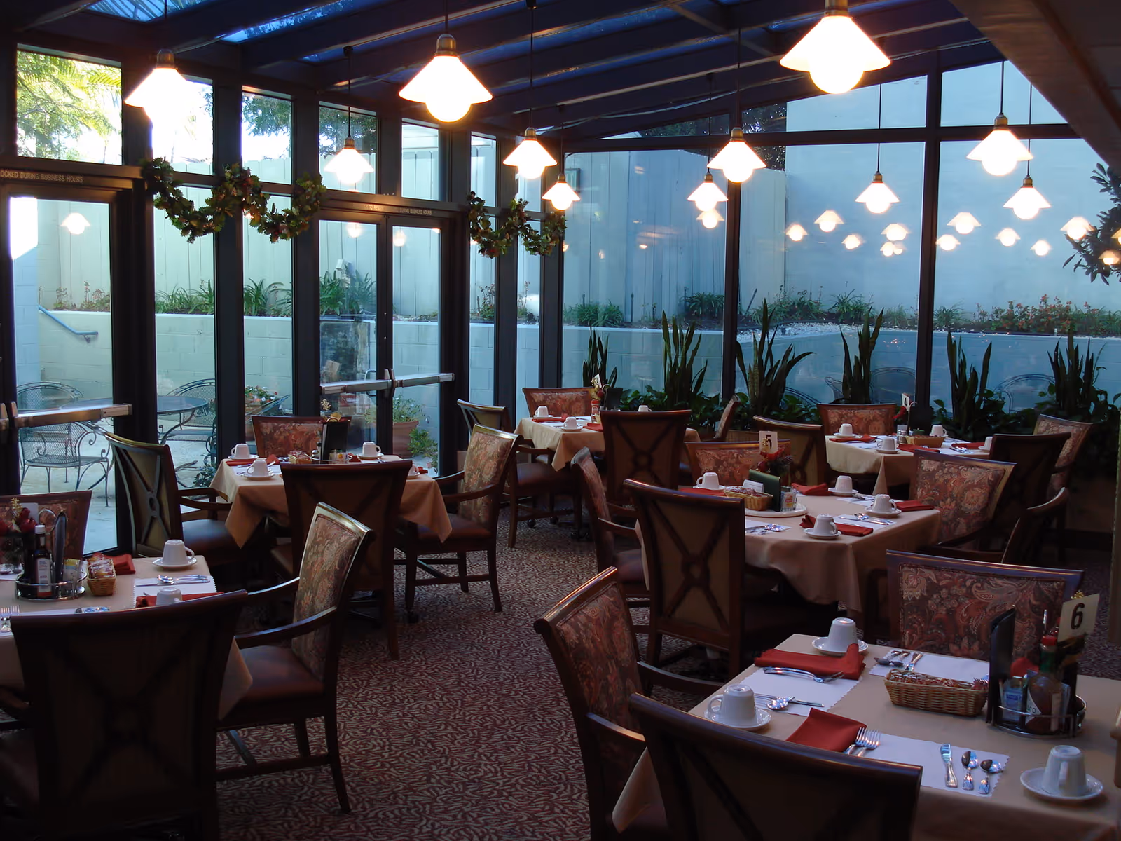 Dining room with multiple tables set with white cups, plates, silverware, and red napkins. The room has large glass windows and doors decorated with green garlands, and there are plants along the walls. Warm pendant lights hang from the ceiling.