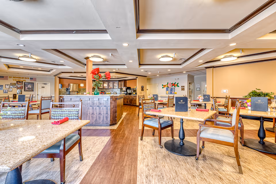 A bright and spacious dining room in an active adult living facility with multiple tables and chairs arranged neatly. Each table has a menu and a red napkin, and there are decorative flowers on some tables. The room features a wooden floor with a mix of light and dark tones, recessed lighting in the ceiling, and a central wooden counter with a vase of red flowers. The walls are painted in warm tones, and there is a bulletin board and patriotic decorations visible in the background.
