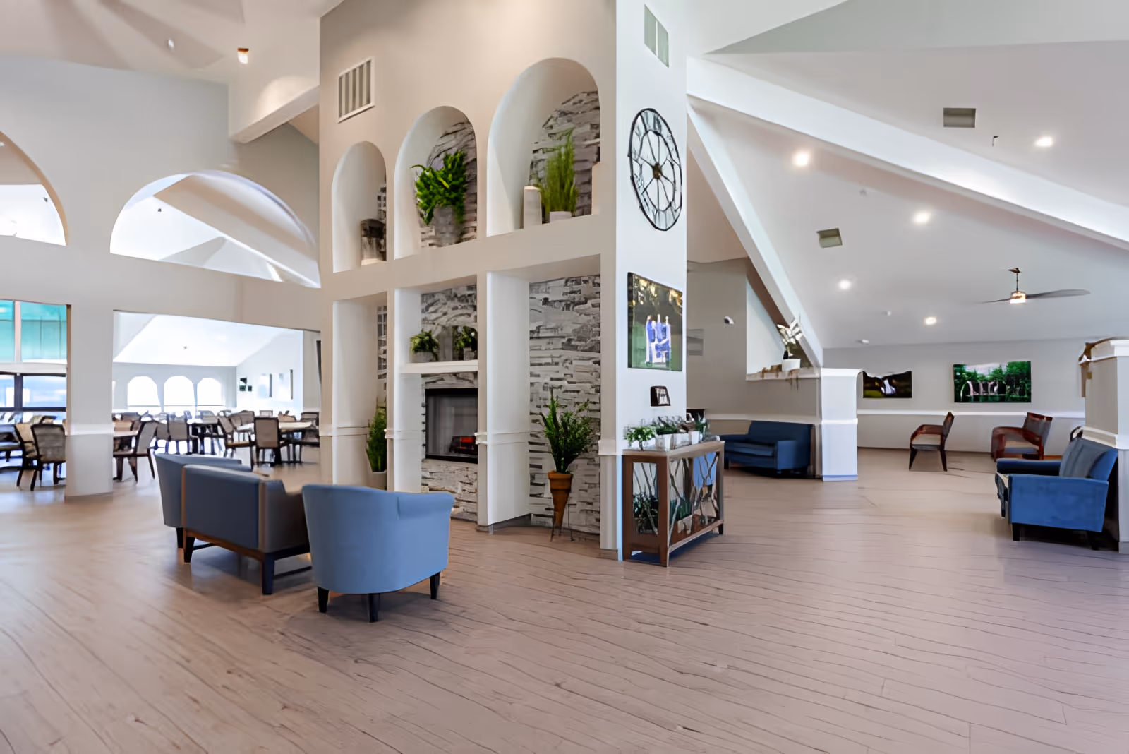 Spacious, bright assisted-living common area with seating clusters, a decorative fireplace, dining tables, and high vaulted ceilings with archways.