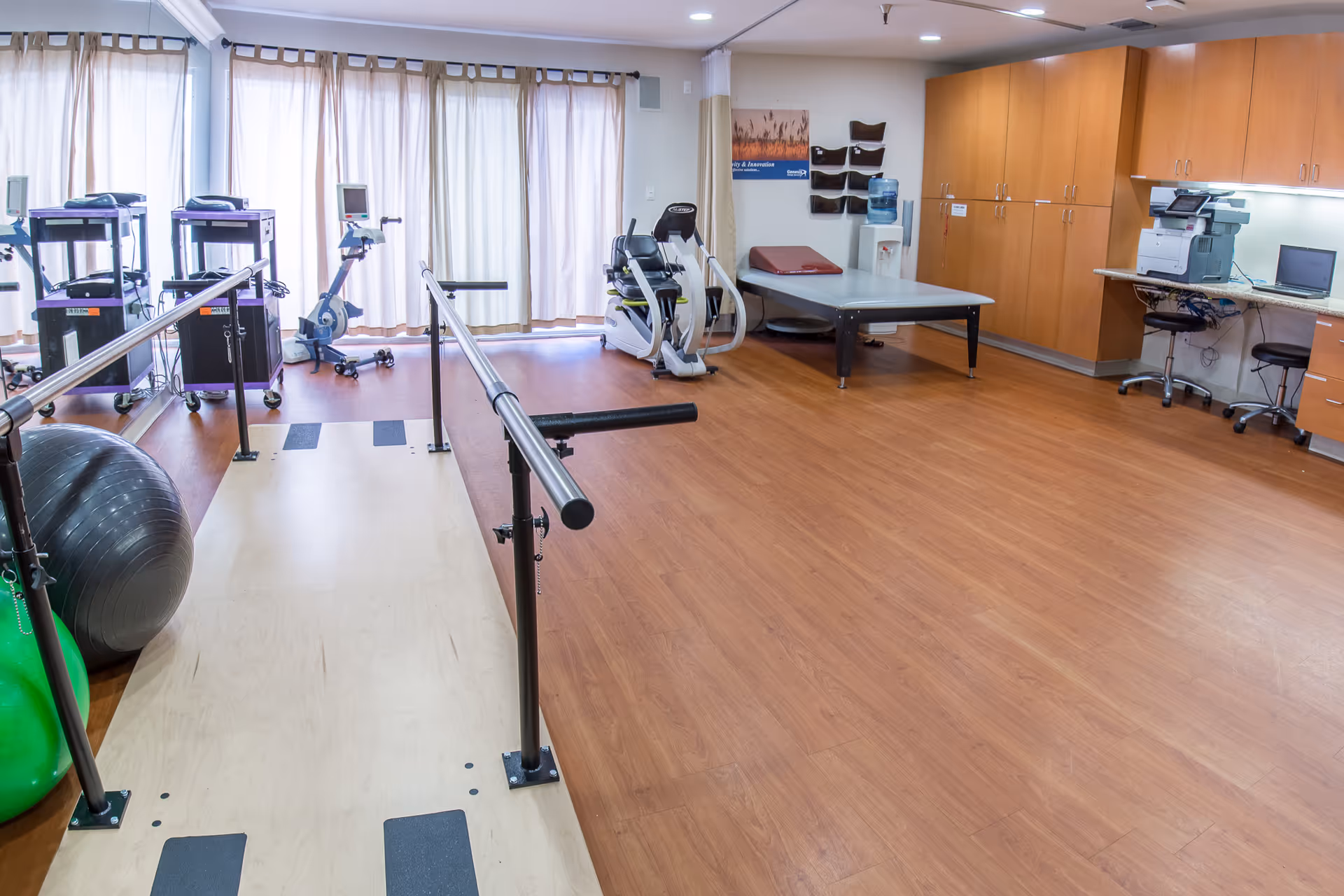 A spacious physical therapy room with wooden flooring, parallel bars for walking exercises, exercise balls, stationary bikes, a treatment table, wooden cabinets, a water dispenser, and a desk with office equipment including a printer and laptop.