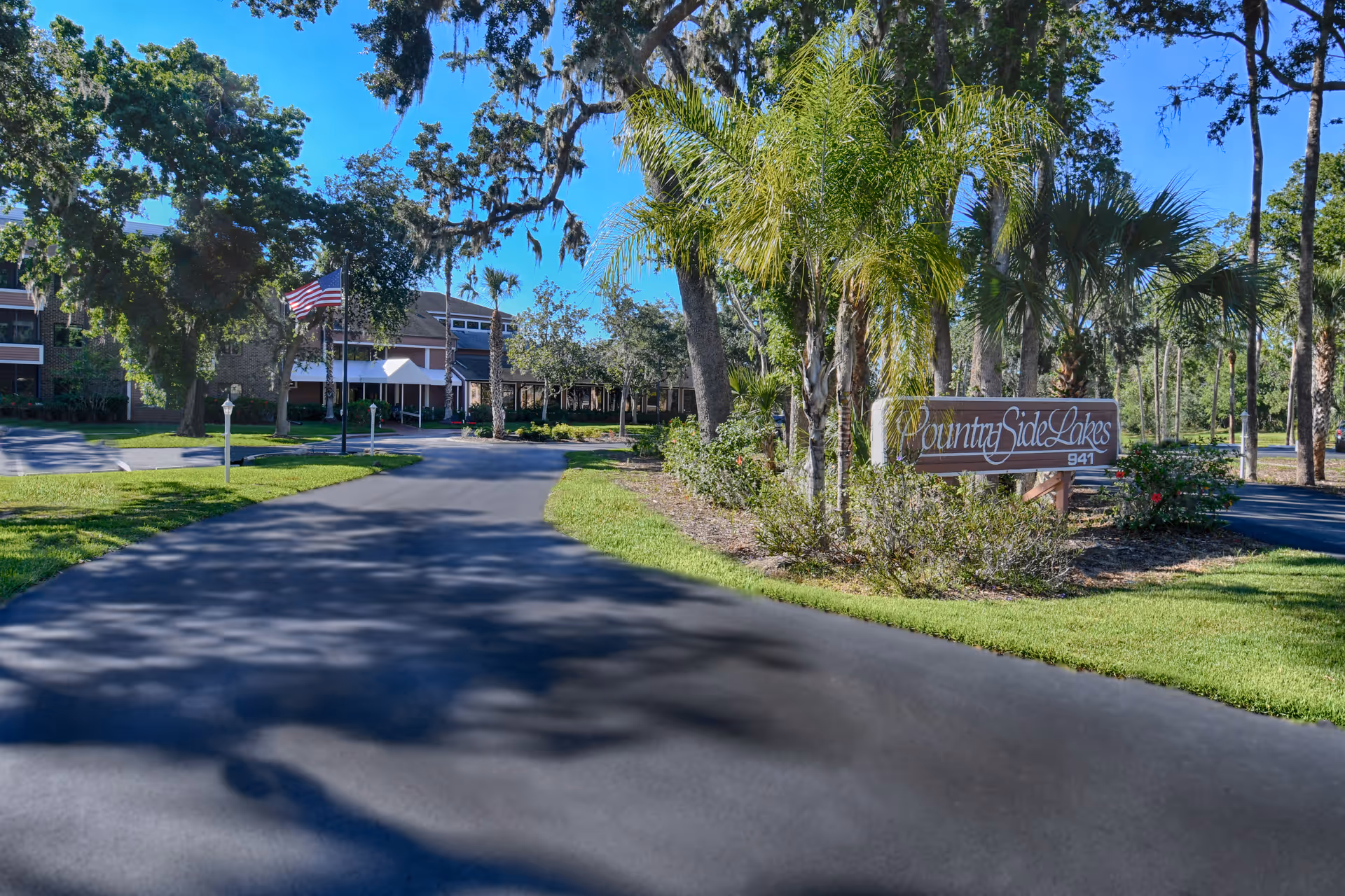 Entrance driveway to CountrySide Lakes senior living facility with a wooden sign displaying the facility name surrounded by palm trees and other greenery. The building is visible in the background with an American flag on a flagpole near the entrance. The sky is clear and blue.