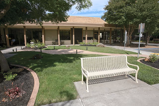 Outdoor area of The Inn on Villa Lane featuring a white metal bench on a concrete path, green grass, landscaped garden beds with plants and trees, and a building with a covered porch in the background under a blue sky with some clouds.