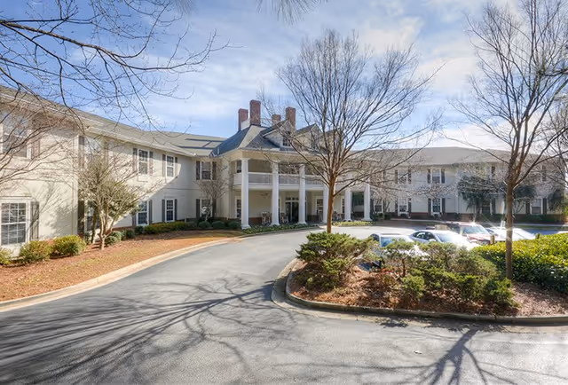 Exterior view of a large two-story senior living facility building with white walls and multiple windows. The entrance features a covered portico with columns. There are several leafless trees and landscaped bushes around the driveway and parking area with a few parked cars. The sky is partly cloudy.