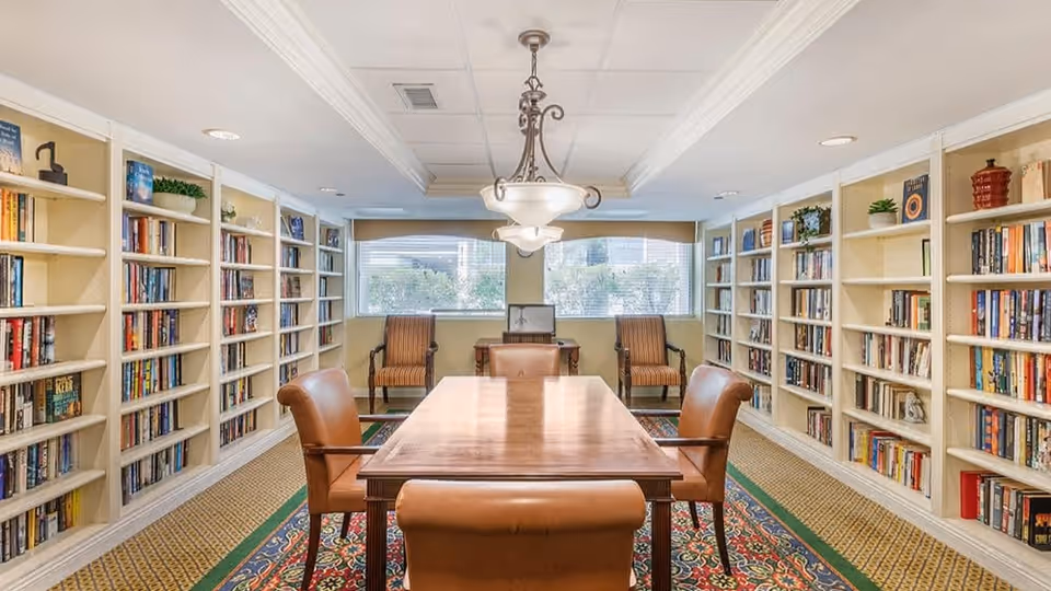 A well-lit room with bookshelves lining both walls filled with books and decorative plants. In the center, there is a wooden table surrounded by four leather chairs. At the far end of the room, two striped armchairs are positioned in front of large windows with a view of greenery outside. A decorative chandelier hangs from the ceiling above the table.