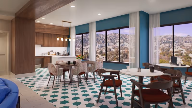 A bright dining area with multiple tables and chairs, featuring large windows with a view of a hillside cityscape. The room has a patterned green and white tiled floor, blue walls, and a kitchen area with wooden cabinets and a hanging light fixture.