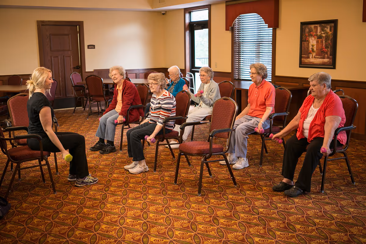 A group of elderly women seated in chairs in a carpeted room participating in a seated exercise class with a female instructor holding small dumbbells.