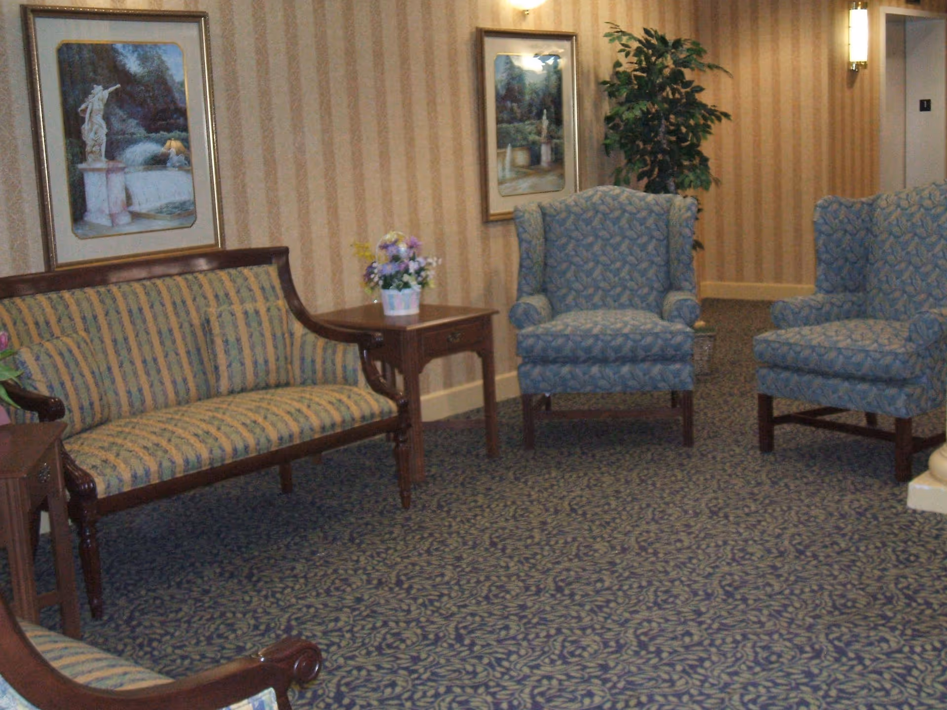 Seating area in a senior living facility with upholstered armchairs, a striped settee, side tables, framed artwork, and a potted plant.