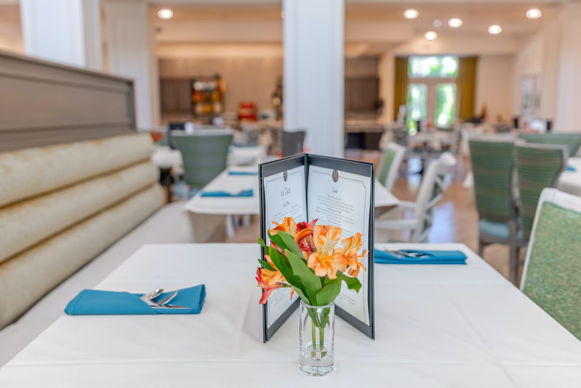 A dining table set with a white tablecloth, a small glass vase with orange and red flowers, a folded menu, and a blue napkin with silverware. The background shows more tables and chairs in a bright dining room with large windows.