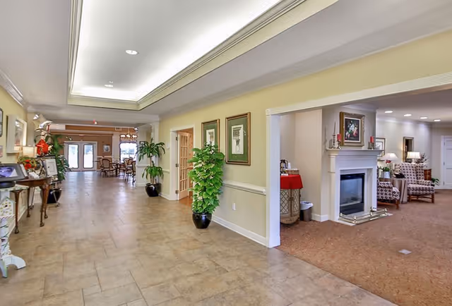 A spacious hallway in a senior living facility with beige tiled flooring and cream-colored walls. The hallway features potted plants, framed artwork, and a side table with decorations. To the right, there is a cozy sitting area with a fireplace, armchairs, and lamps. In the background, a dining area with tables and chairs is visible near large windows.