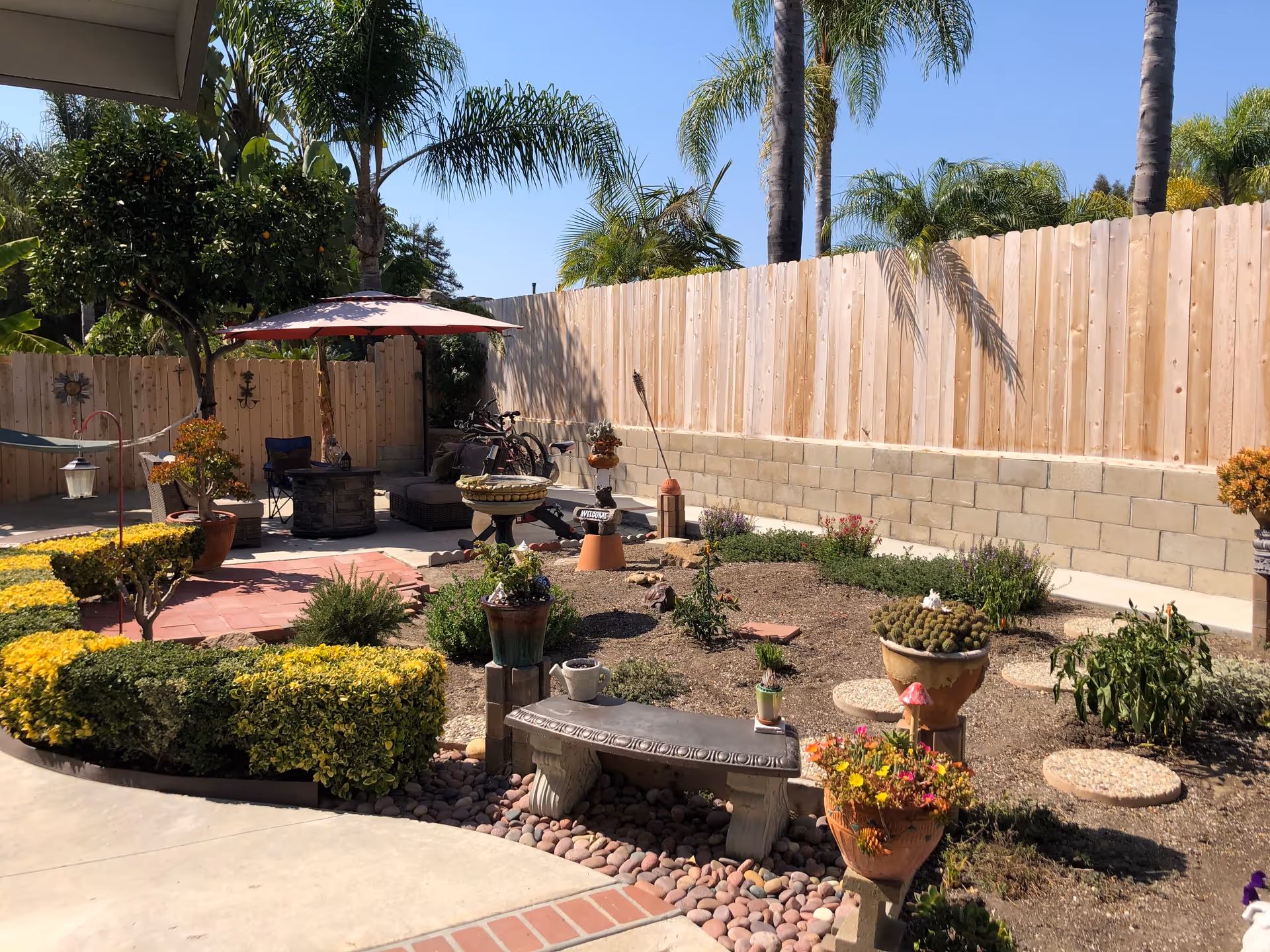 A sunny outdoor garden area with various plants, shrubs, and potted flowers. There is a stone bench in the foreground, a patio umbrella shading a seating area with chairs, and a wooden fence in the background. Palm trees are visible beyond the fence under a clear blue sky.