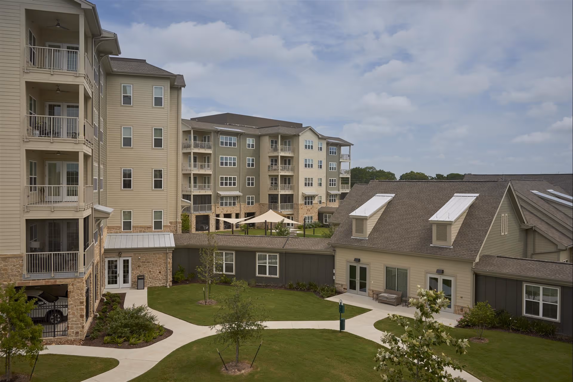 Exterior view of The Langford Methodist Retirement Community showing multiple beige and green buildings with balconies, a well-maintained lawn with small trees, paved walkways, and a covered outdoor seating area under beige shade sails.