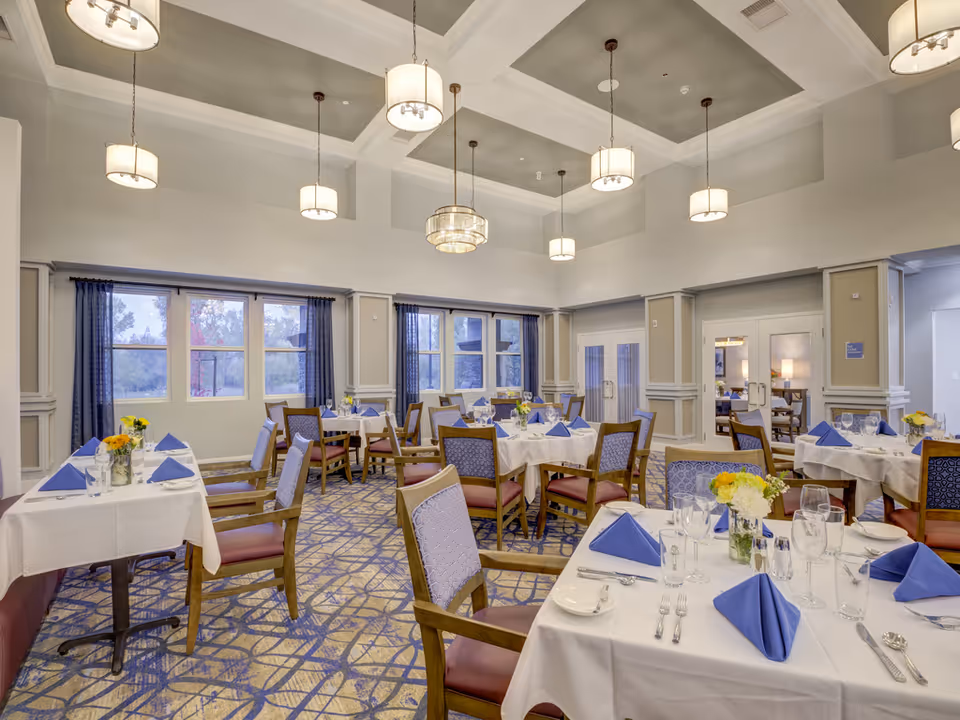 Well-lit formal dining room with tables covered in white linens, blue napkins, floral centerpieces, and hanging pendant lights.
