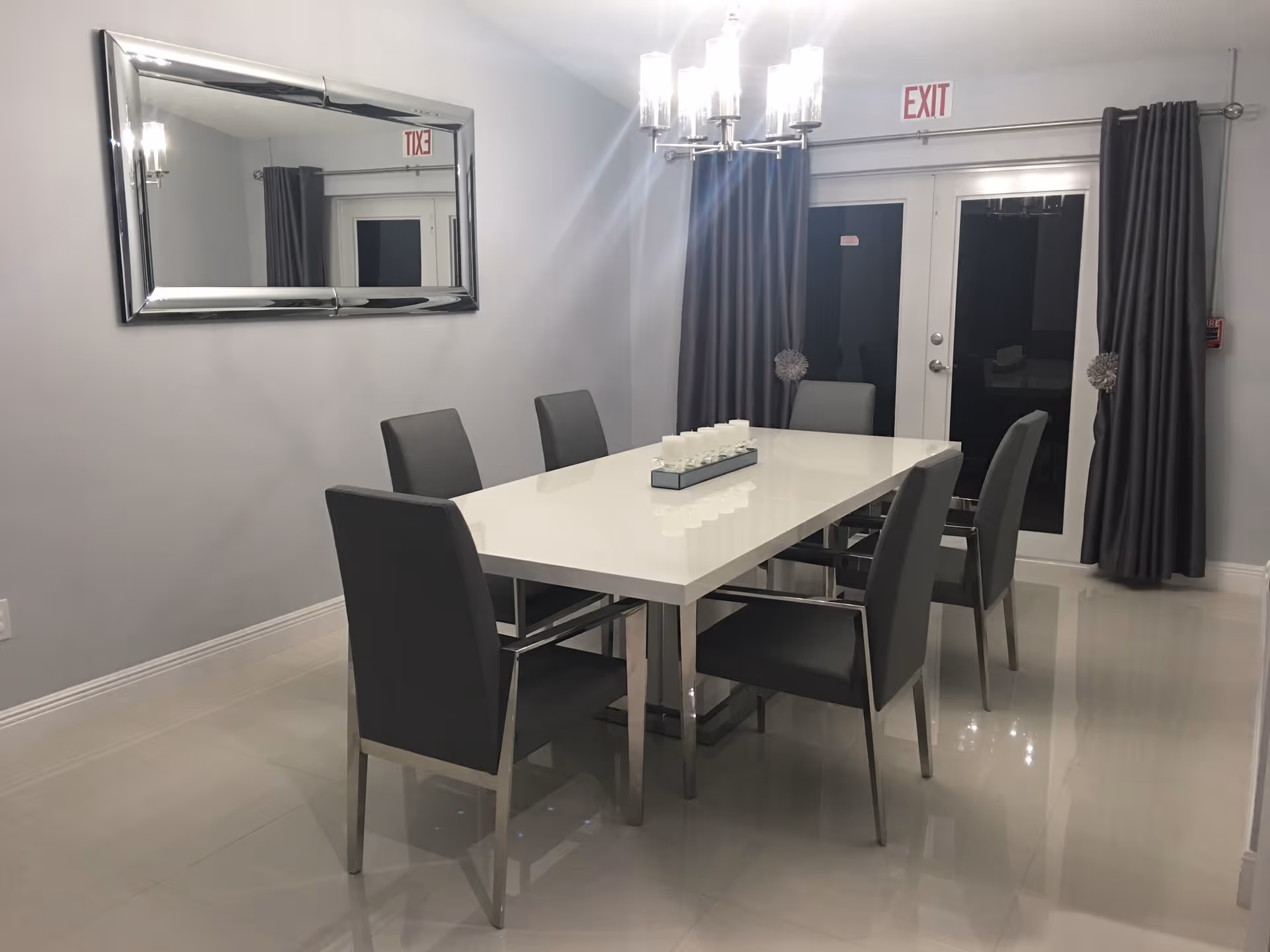 A modern dining room with a white rectangular table surrounded by six gray chairs. A decorative candle holder with multiple candles is centered on the table. The room features a large rectangular mirror on the left wall, gray curtains covering glass doors at the back, and a chandelier hanging above the table. An exit sign is visible above the doors.