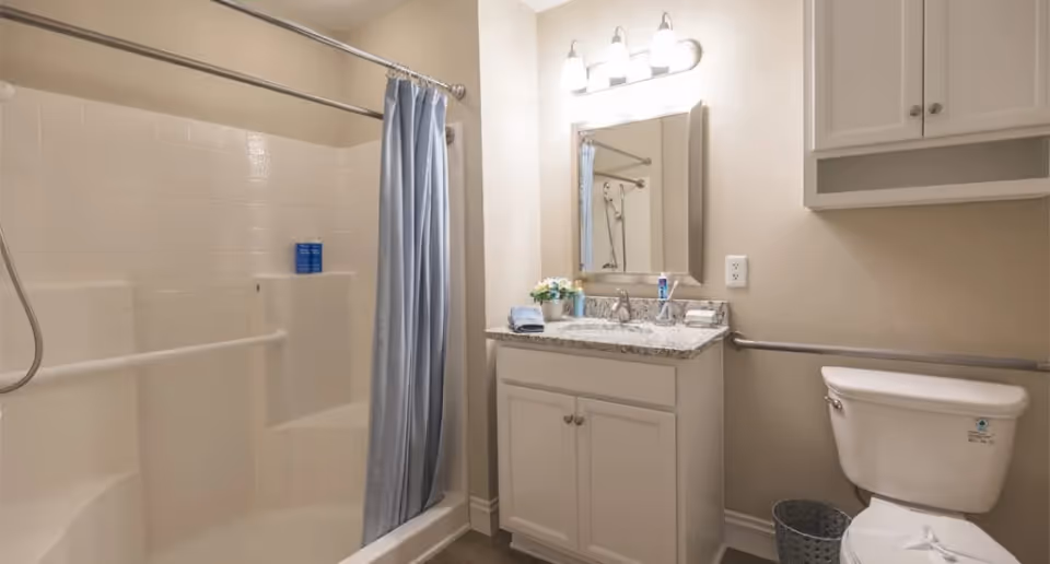 Accessible bathroom with a shower-tub and blue curtain, vanity with mirror and granite countertop, toilet and grab bars.