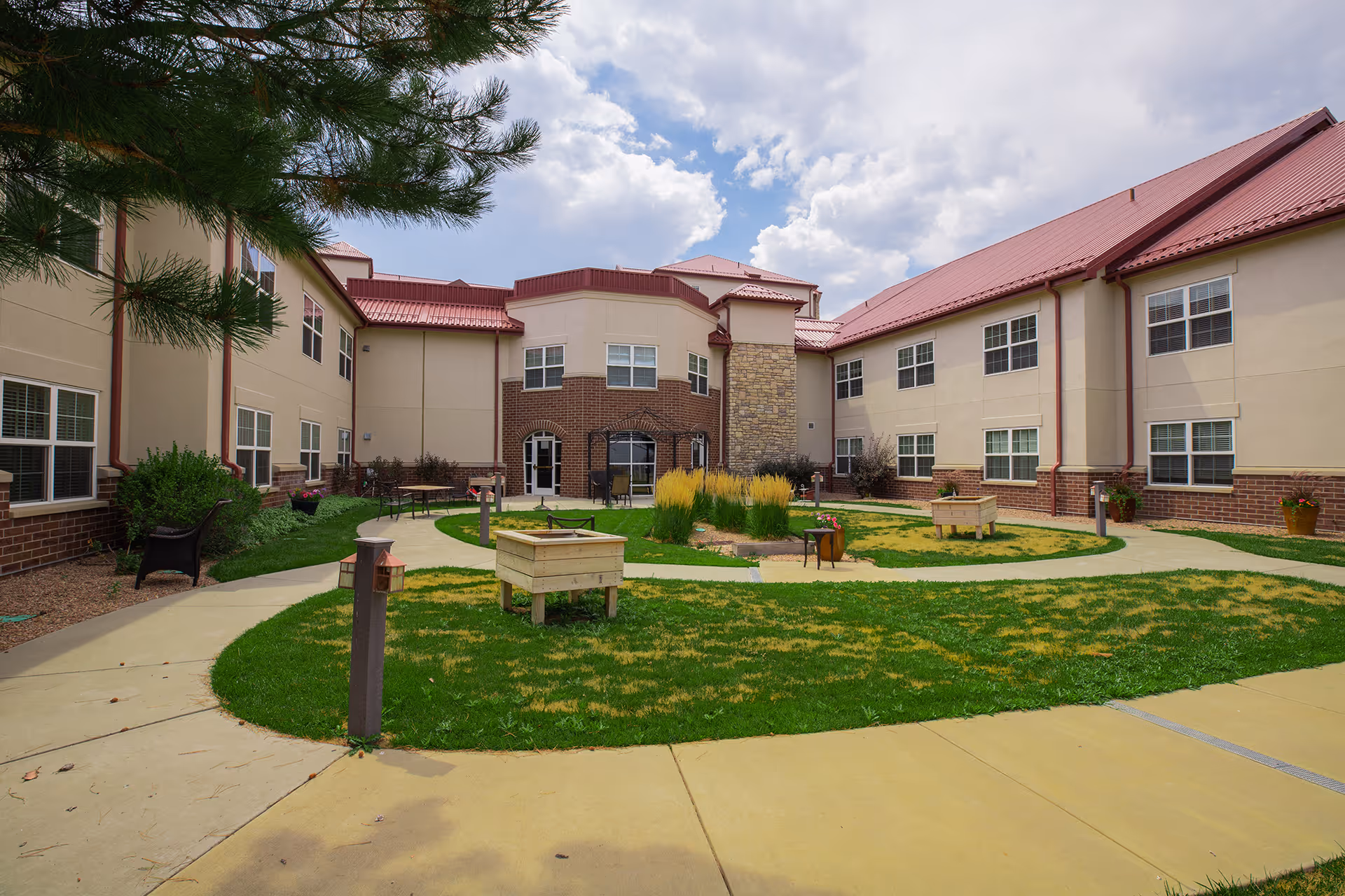 Outdoor courtyard area of a senior living facility with green grass, paved walkways, benches, planters, and a two-story building with red roof and beige walls surrounding the courtyard under a partly cloudy sky.
