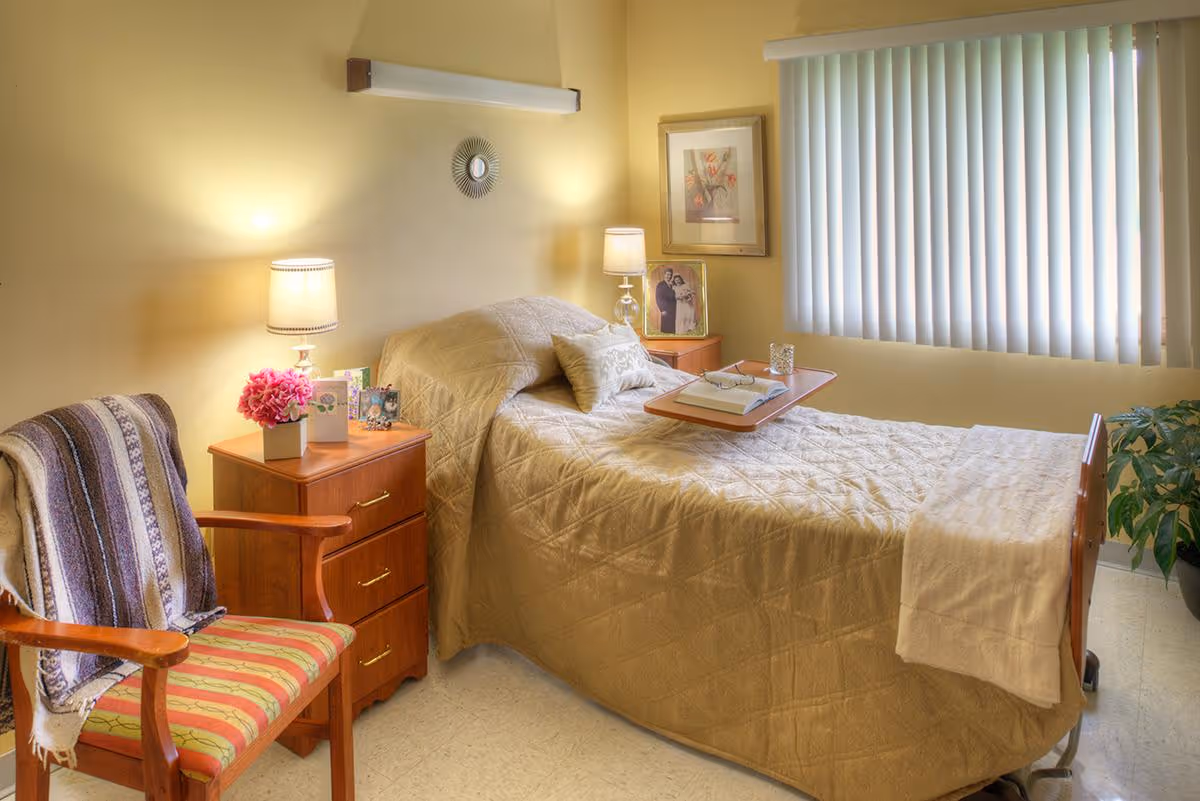 A neatly made single bed with bedside tables, lamps, a chair, and a window with vertical blinds in a warmly lit room.