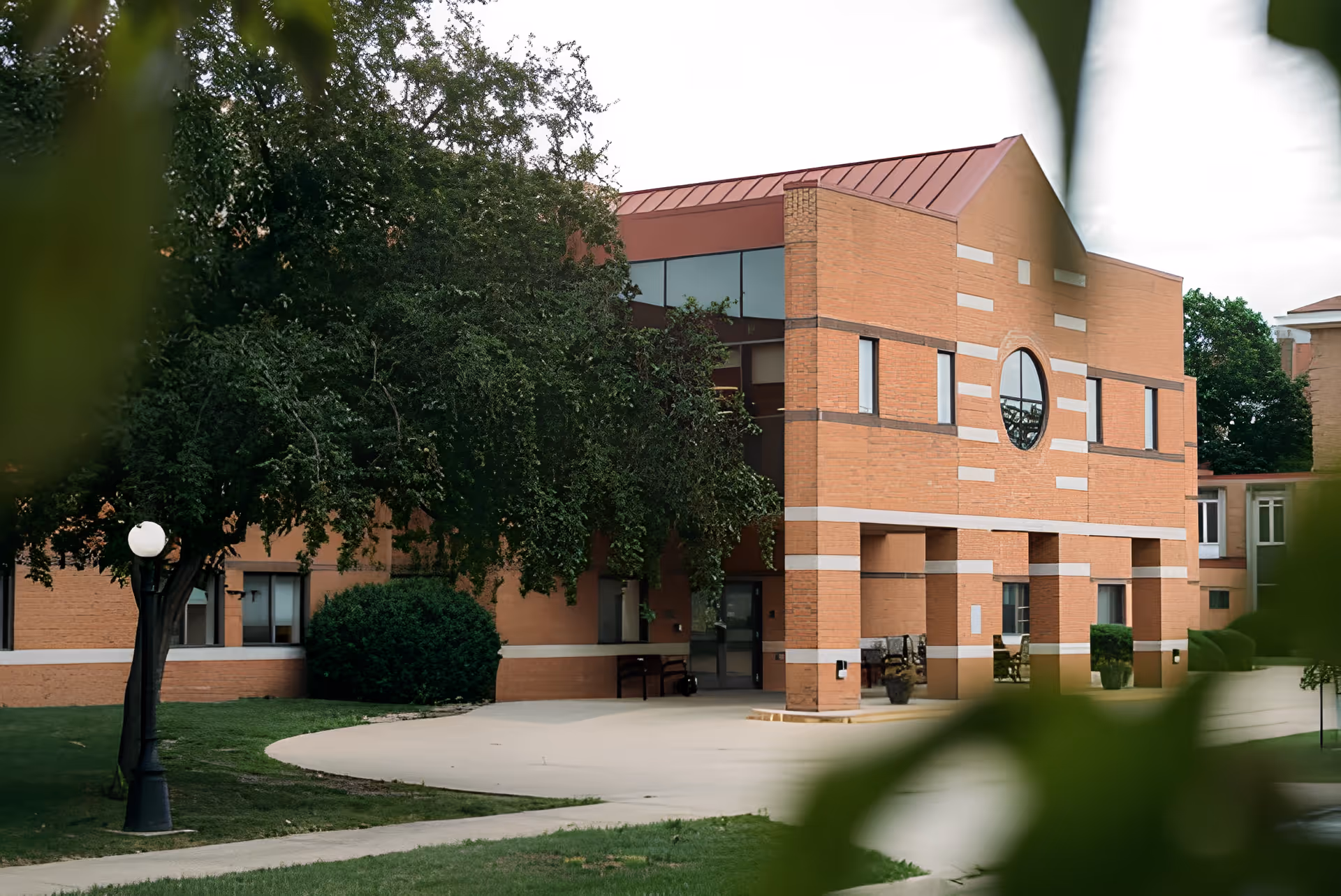 Exterior view of a brick building with a red roof and a circular window, partially obscured by green trees and foliage. There is a paved walkway and a black lamp post in the foreground.