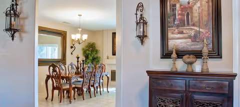 View through an entryway into a dining room with a wooden dining table and six matching chairs. The room is decorated with a large mirror on the left wall, a green potted plant in the corner, and a chandelier hanging above the table. On the right side of the entryway, there is a wooden cabinet with decorative items and a framed painting above it. The walls are light-colored, and the floor is tiled.