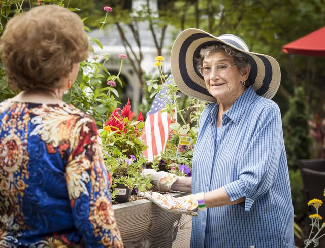 Two elderly women are gardening outdoors. One woman, wearing a large striped sunhat, glasses, a blue striped shirt, and gardening gloves, is tending to a raised garden bed filled with colorful flowers and an American flag. The other woman, seen from behind, is wearing a colorful patterned blouse. Trees and outdoor furniture are visible in the background.