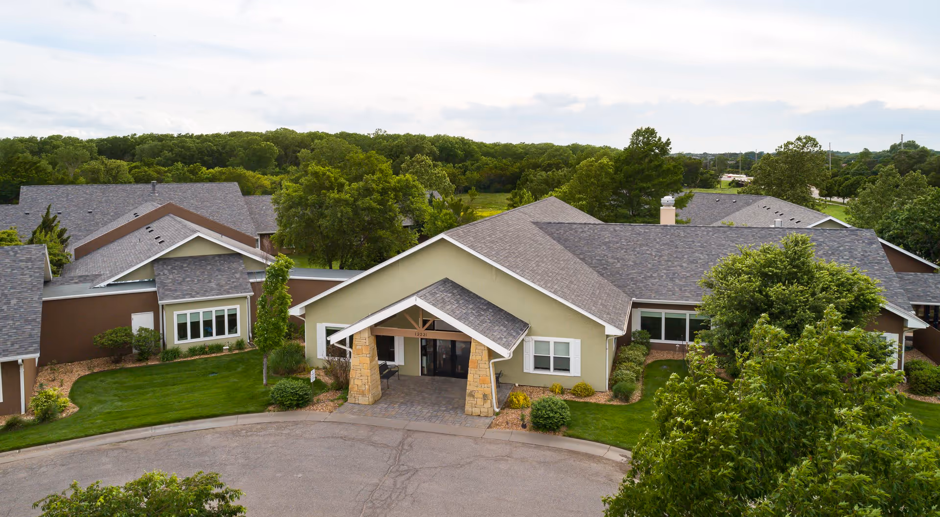 Aerial view of Homestead Assisted Living & Memory Care of Wichita showing the front entrance of a single-story building with a covered porch supported by stone pillars, surrounded by green trees and well-maintained landscaping.