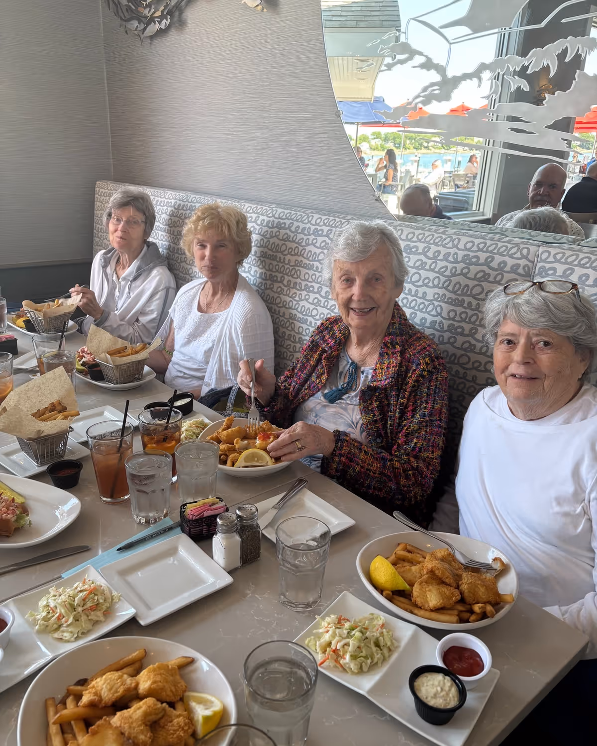 Four elderly women sitting at a restaurant table enjoying a meal together. The table is set with plates of fried fish, French fries, coleslaw, and various drinks. Behind them is a patterned cushioned bench and a large mirror reflecting the outdoor seating area with umbrellas and people.