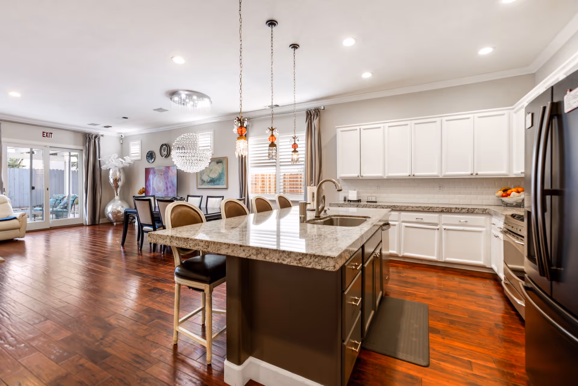 A spacious and modern kitchen and dining area with wooden floors. The kitchen features a large island with a granite countertop, a sink, and several bar stools. White cabinets line the walls, and stainless steel appliances are visible. The dining area has a table with chairs and a large decorative light fixture above it. Large windows and glass doors provide natural light and a view of an outdoor seating area.