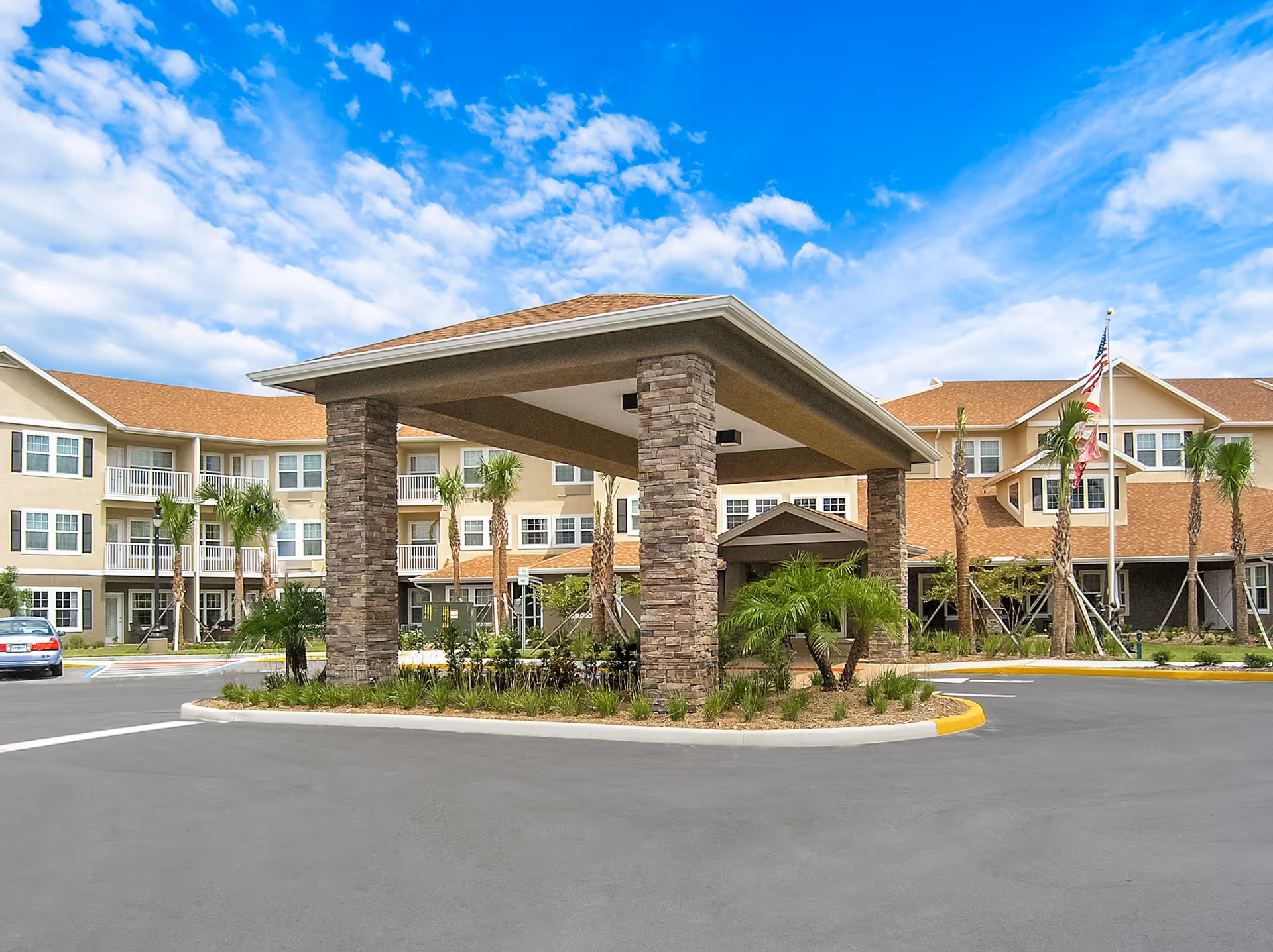 Exterior view of a senior living facility with a covered entrance supported by stone pillars, surrounded by palm trees and landscaping under a partly cloudy blue sky.