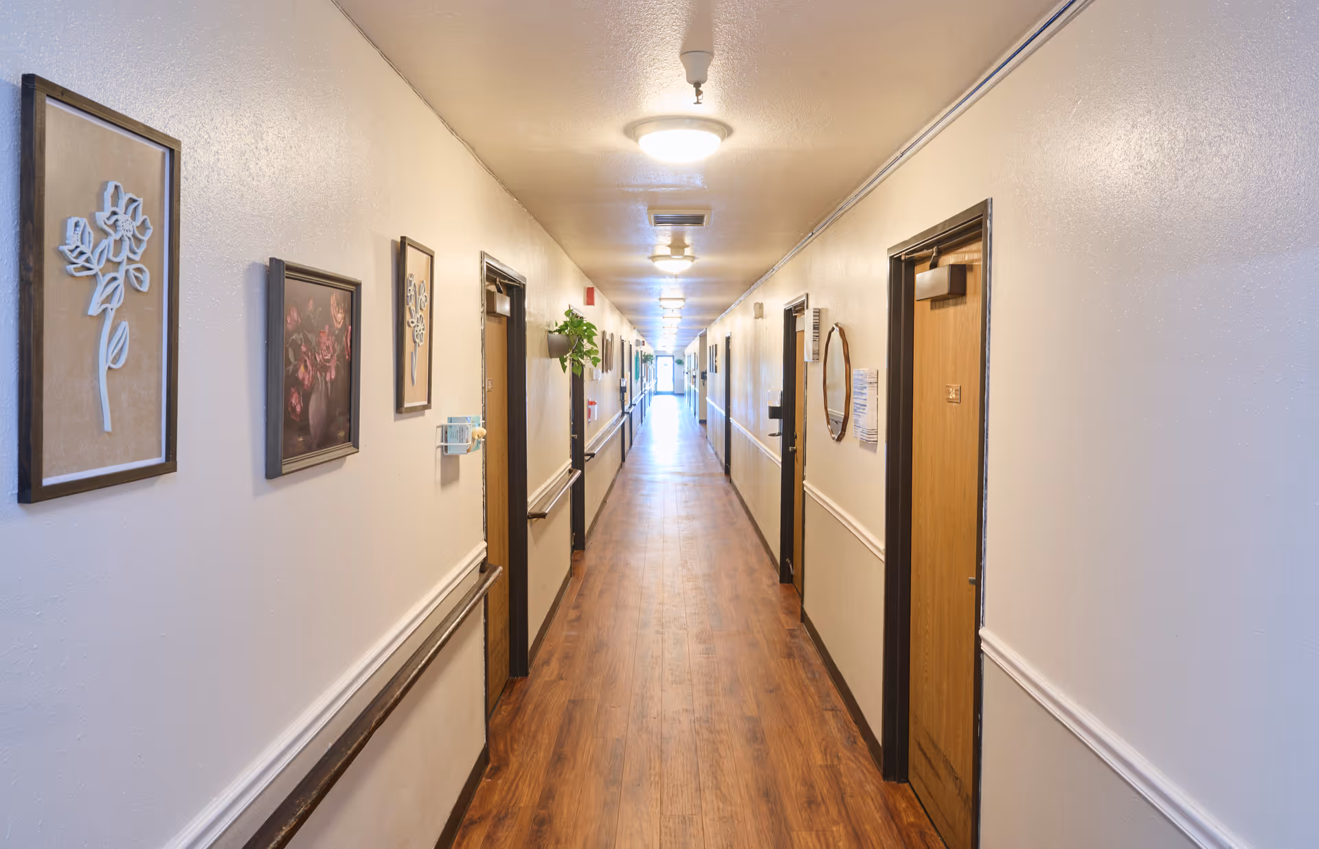 A long, well-lit hallway in a residential facility with wooden doors on both sides, light-colored walls decorated with framed artwork and a small plant, and wooden flooring.